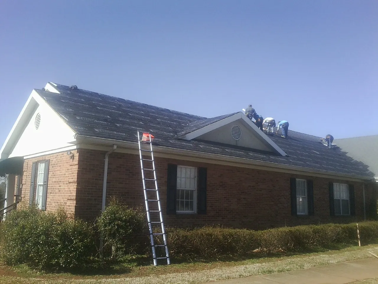 Crew tearing shingles off a brick ranch roof with a ladder set — Emergency Roof Repair work by our crew in Fort Rock