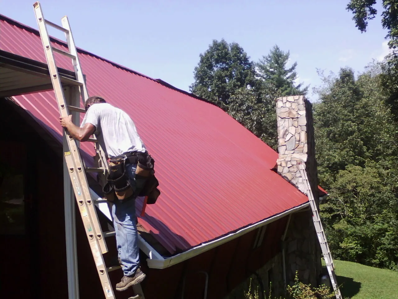 Roofer on a ladder beside a red metal roof and stone chimney — Soffit & Fascia Repair by Roofing Brothers in Fort Rock